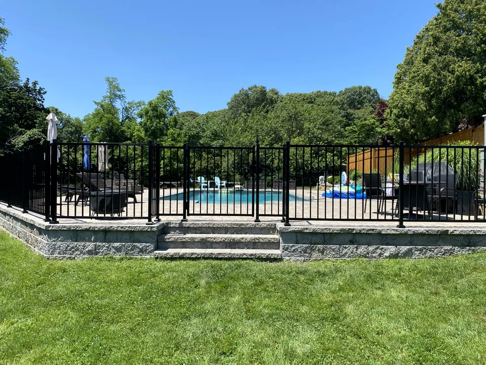Four-foot black aluminum pool fence installed along a raised retaining-wall and patio structure in Miller Place, New York.