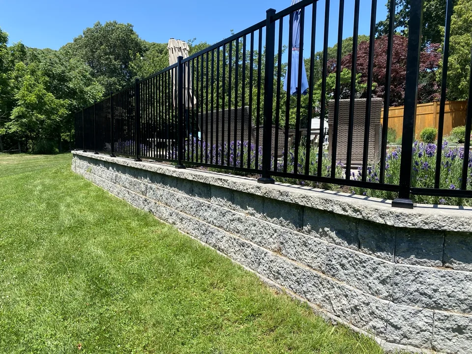 Four-foot black aluminum fence following the top of a raised retaining wall beside a pool patio in Miller Place, New York.