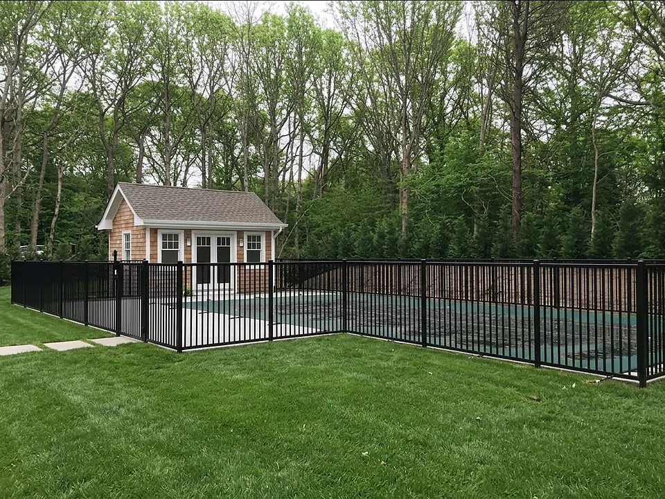 Black aluminum pool fence enclosing an inground pool beside a cedar-shingled poolhouse, backed by an evergreen screen and woodland, in East Hampton, New York