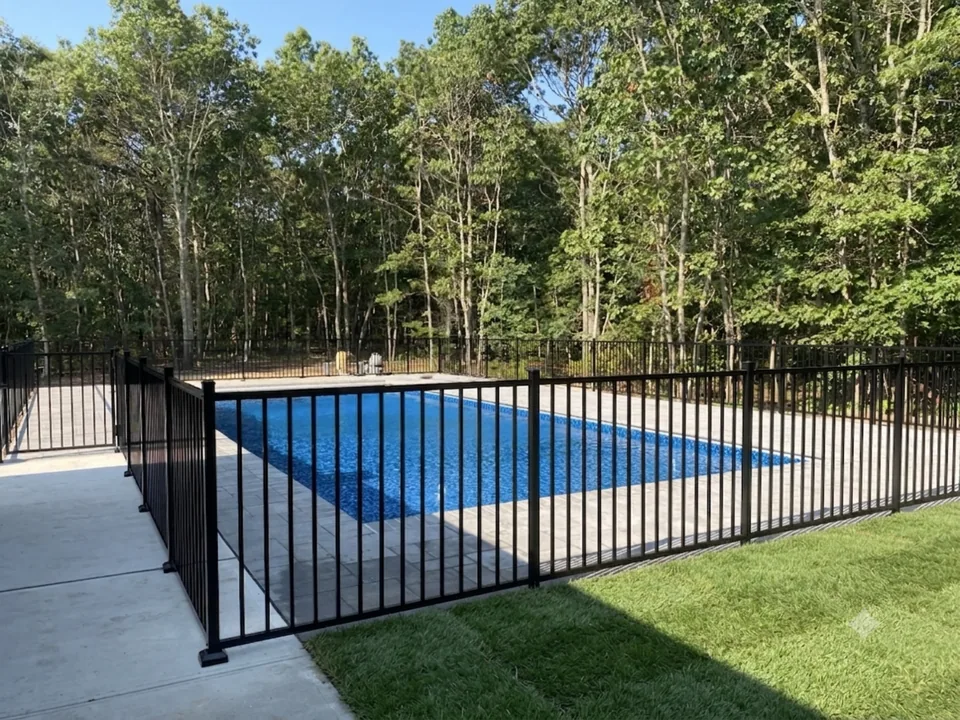 Four-foot black aluminum fence enclosing a rectangular inground pool on a paver deck surrounded by woods in Manorville, New York
