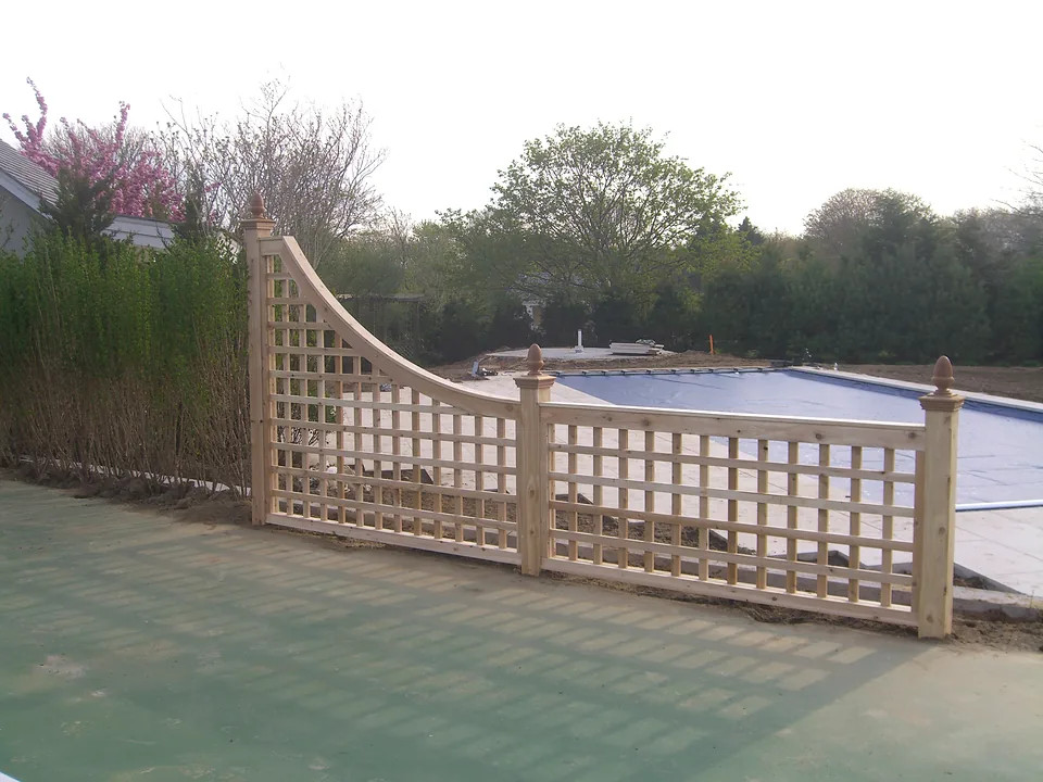 Cedar lattice accent panel with a swept curved top rail standing between a tennis court and a pool terrace in Southampton, New York