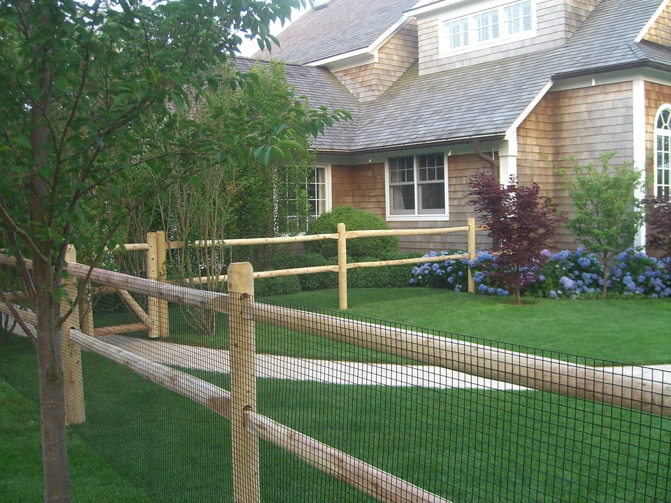 Peeled cedar post and rail fence with black welded wire backing running across a lawn beside a shingled house and hydrangeas in Southampton, New York