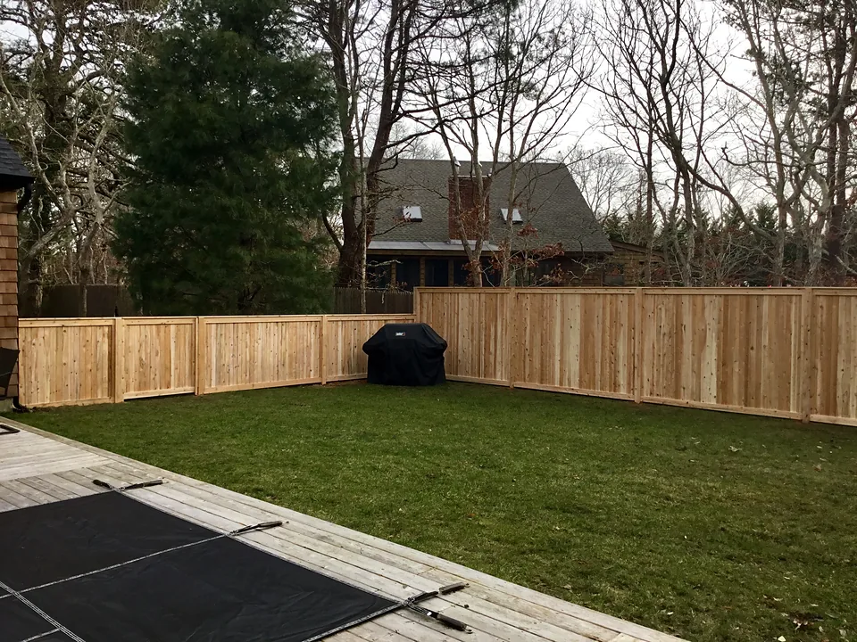 Corner of a natural cedar tongue-and-groove privacy fence enclosing a lawn beside a pool deck, four-foot and six-foot runs meeting at the property line, in East Hampton, New York