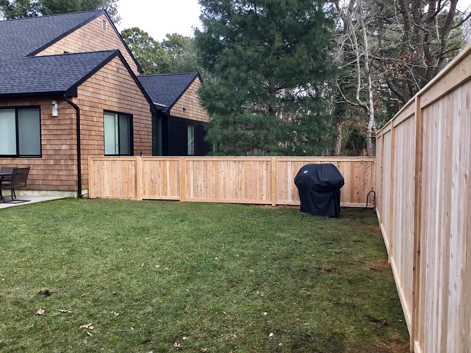 Natural cedar tongue-and-groove privacy fence, a four-foot run along a cedar-shingled house stepping up to a six-foot run at the property line, in East Hampton, New York