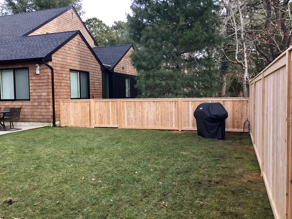 Natural cedar tongue-and-groove privacy fence running across a back lawn, the four-foot run stepping up to six feet at the corner, beside a cedar-shingled house in East Hampton, New York