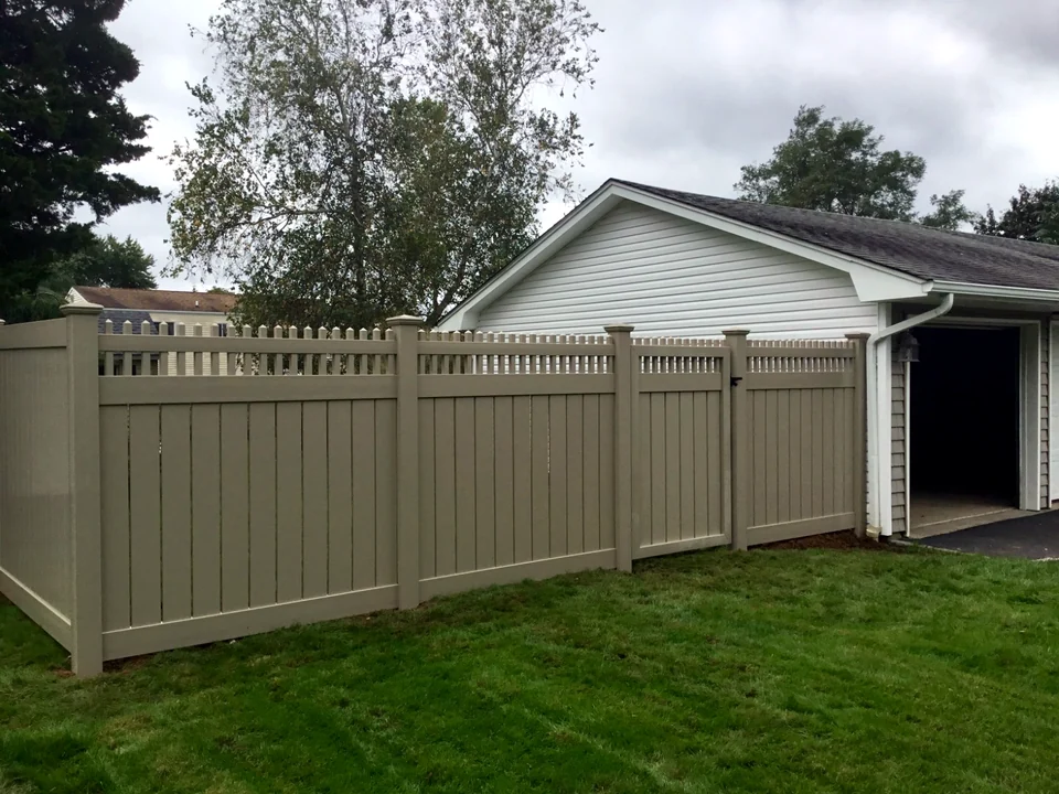 Angled side-yard view of tan PVC privacy fencing with a decorative picket top in Coram, New York.