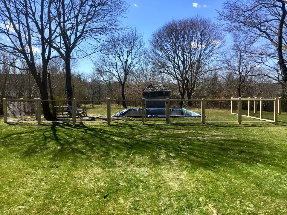 Full-yard view showing the cedar framed-wire pool enclosure within the mature landscape in Center Moriches, New York.