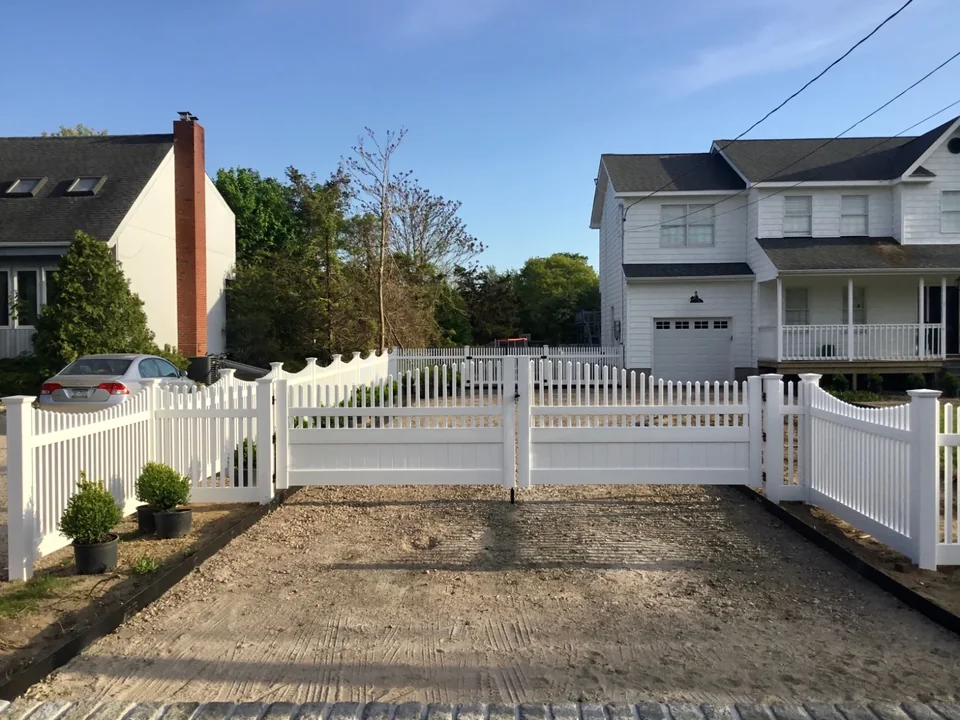 Wide job-site view of a custom white PVC fence and driveway gate project in Hampton Bays, New York.