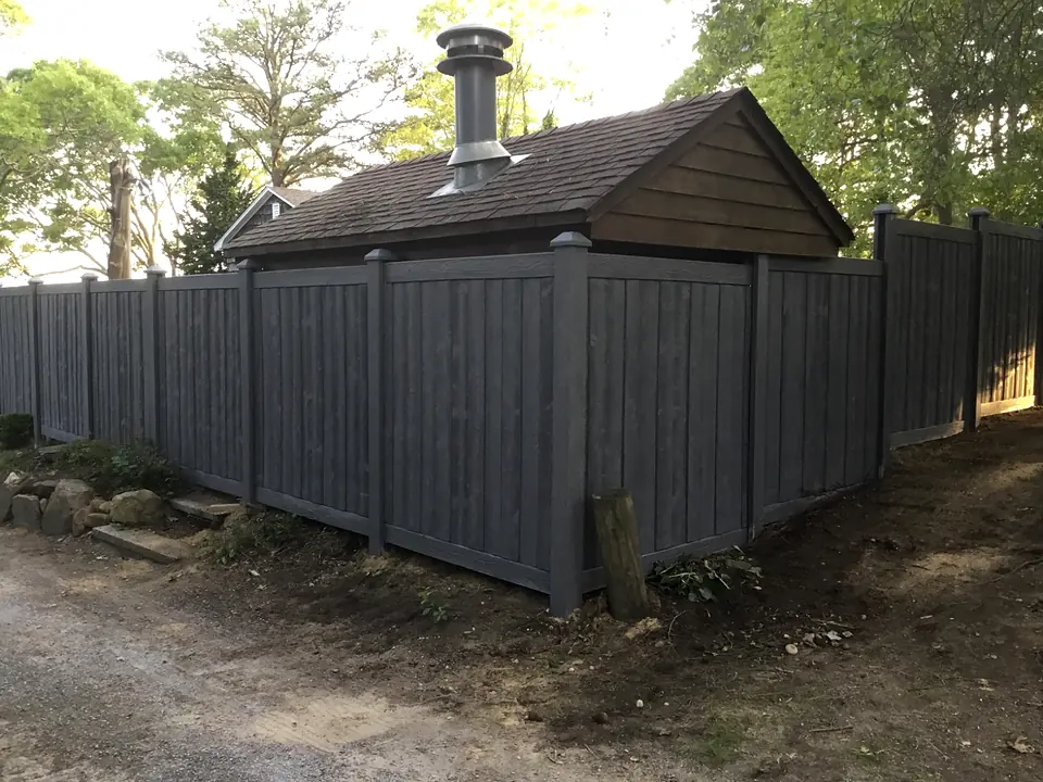 Gray wood-look molded privacy fence beside a cedar-shingled outbuilding