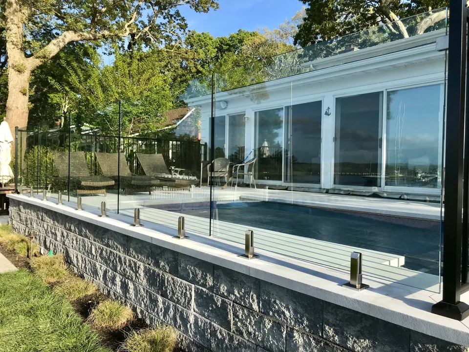 Frameless glass pool fence wrapping the corner of a raised stone patio in Southold, New York.